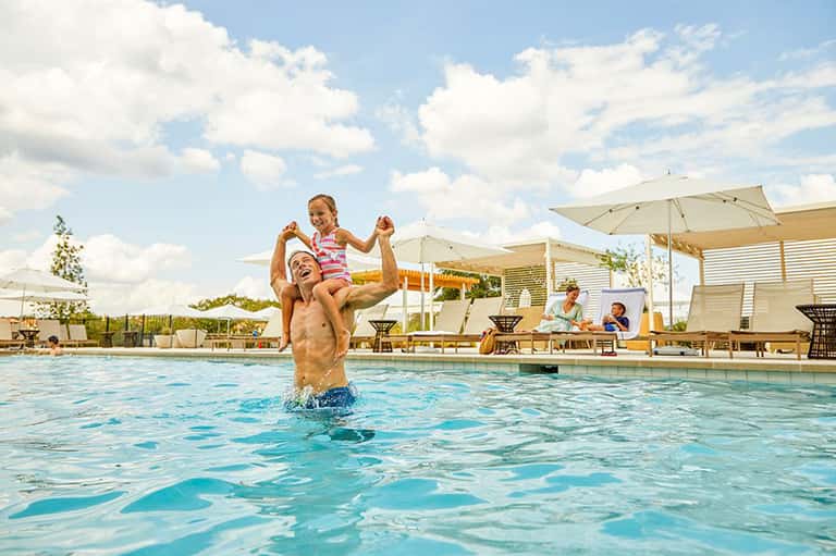 Father lifting child in resort pool with umbrellas and lounge seating in background
