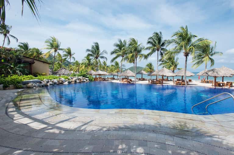 Tropical resort pool with tiki umbrellas, lounge chairs, palm trees, and ocean backdrop