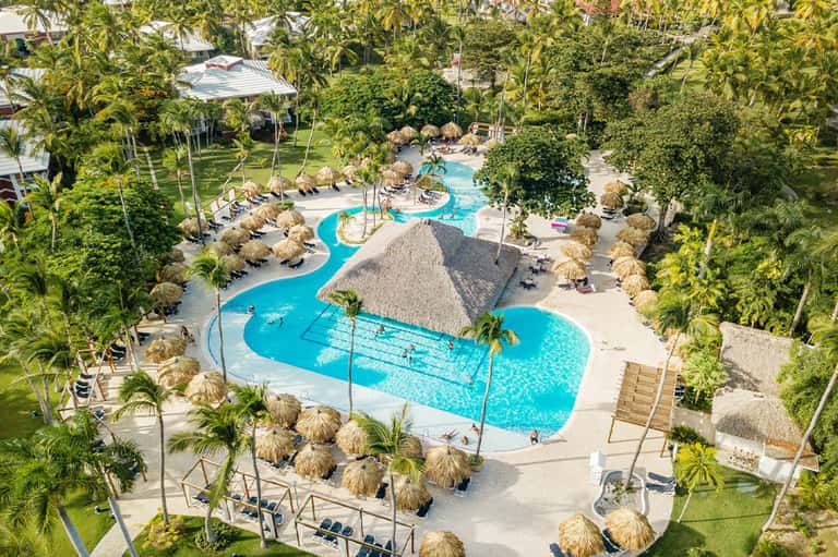 Aerial view of turquoise lagoon pool surrounded by tiki umbrellas, palm trees, and lush tropical grounds