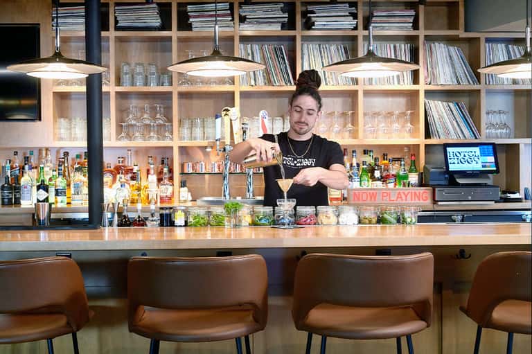 Craft cocktail bar with bartender mixing drinks, wooden shelving with spirits, and warm industrial lighting