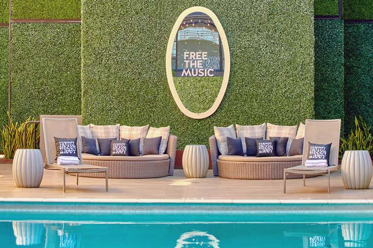 Resort pool deck with green artificial grass wall, rattan seating, and neon oval sign reading 'Free the Music'
