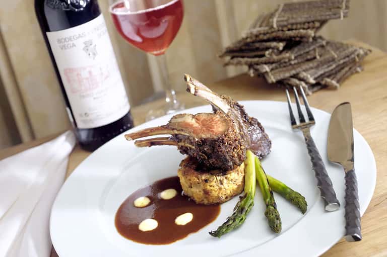 Plated lamb chop dish with wine glass and bottle on wooden table with garnish and sauce