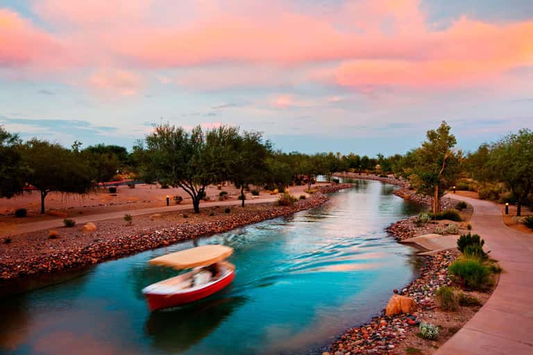Scenic river at sunset with boats, trees, and pink sand landscape under colorful sky