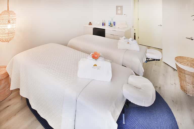 Spa treatment room with two massage tables and amenities counter with orange flower accent