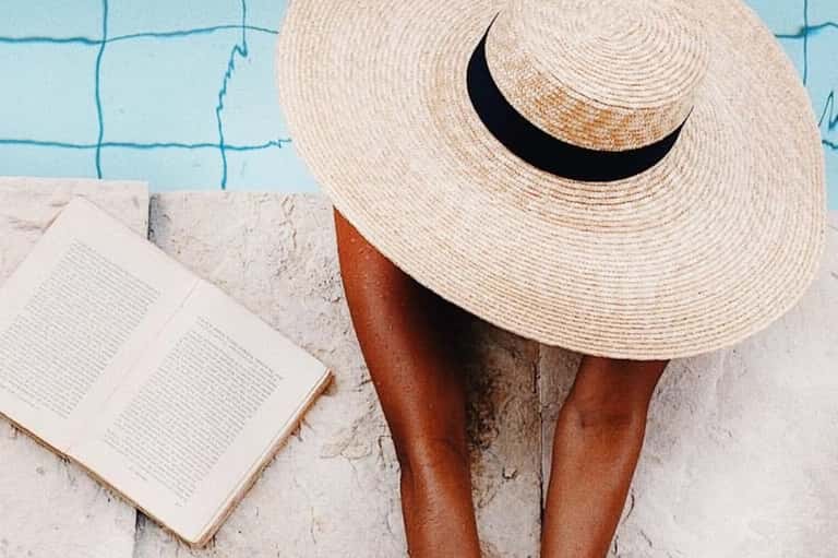 Person poolside wearing oversized straw hat, reading book by turquoise water