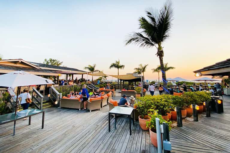 Sunset rooftop lounge with coral furniture, palm trees, and mountain views with guests relaxing