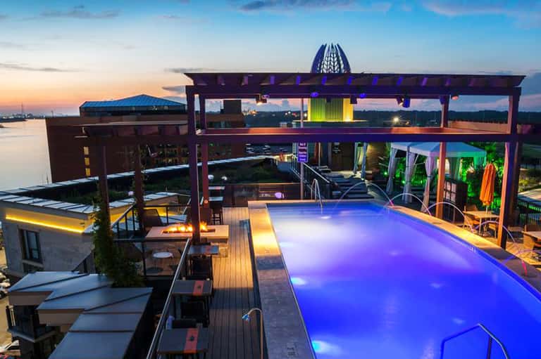 Rooftop infinity pool glowing blue at twilight with lounge chairs, fire pit, and waterfront landscape views
