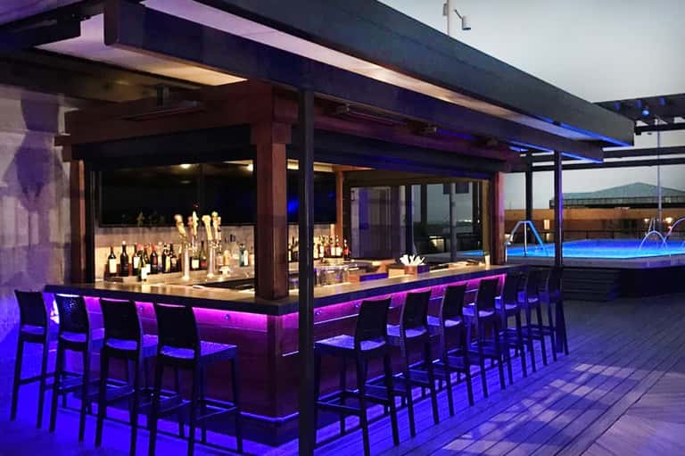 Modern rooftop bar with illuminated purple accent lighting, liquor display, and ocean views at dusk