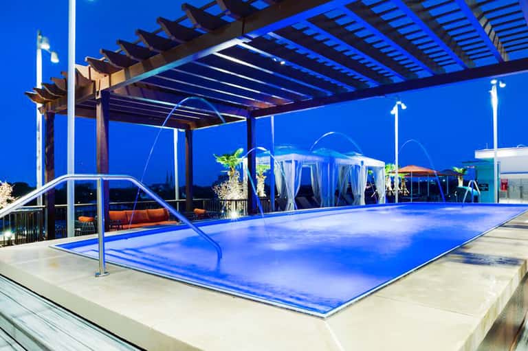 Modern rooftop pool with bright blue water, pergola, lounge seating, and evening city lights