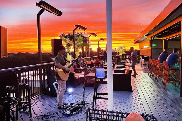 Live musician performing on a rooftop terrace at golden sunset with palm trees and city skyline behind