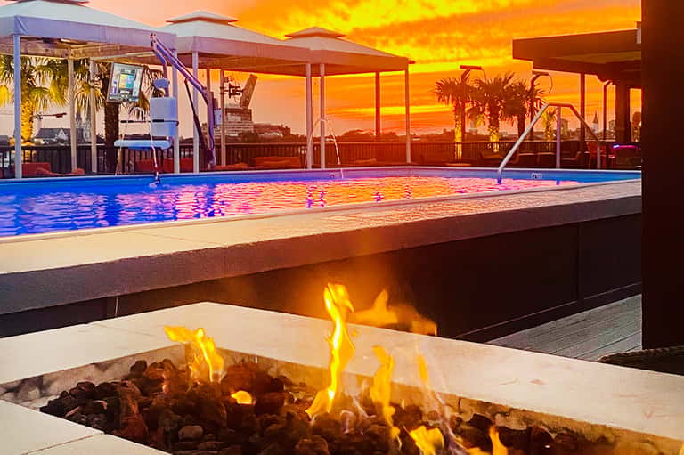 Resort pool at sunset with fire pit, palm trees, and colorful sky reflection