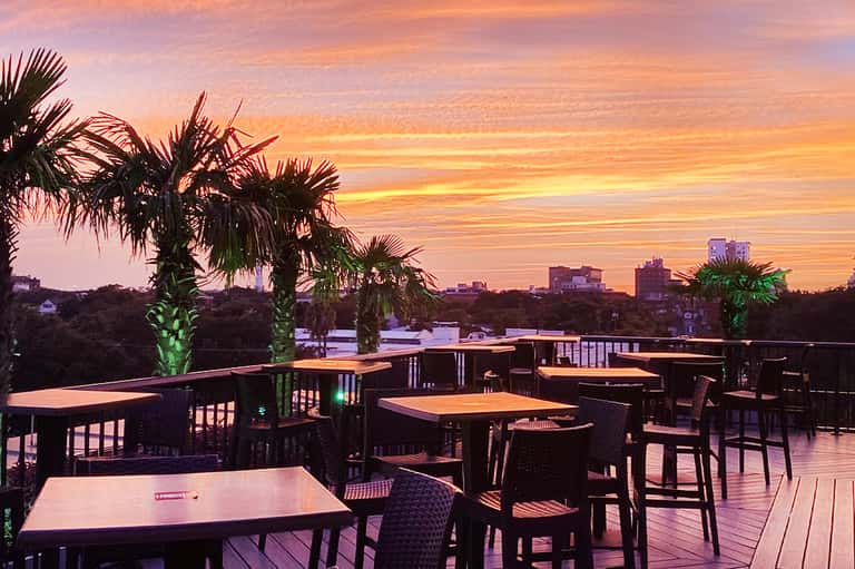 Outdoor rooftop dining area at sunset with palm trees, wooden tables, and colorful sky reflection