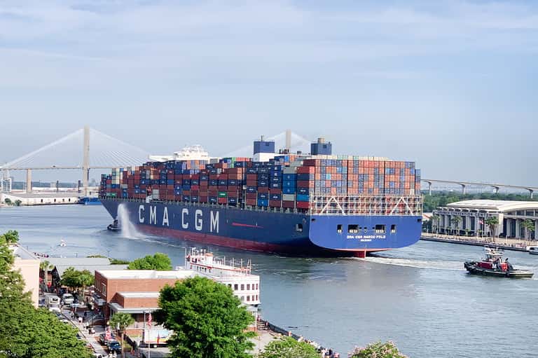 Large cargo ship with stacked shipping containers passing under bridge near waterfront city