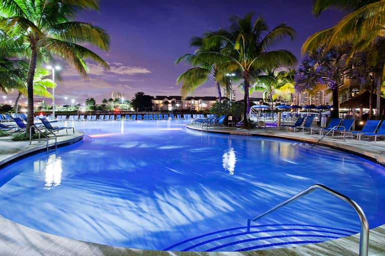 Illuminated resort pool at dusk with palm trees and blue lounge chairs