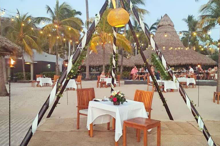 Beach bonfire dinner setup with tiki torches, white tablecloth, and palm surroundings