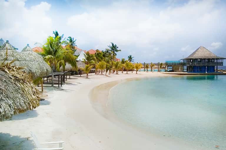 Tropical beach resort with white sand, turquoise water, thatched umbrellas, palm trees, and beachfront buildings