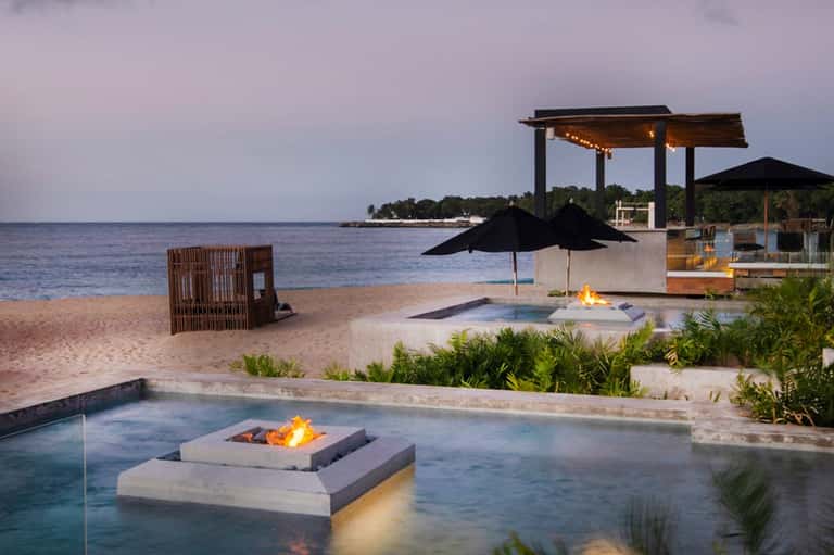 Beachfront fire pit lounge at dusk with modern pergola, fire feature, and ocean views with resort buildings.