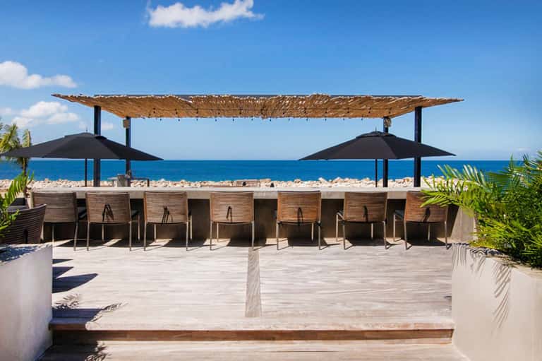 Beachfront dining area under woven roof with umbrellas overlooking turquoise ocean