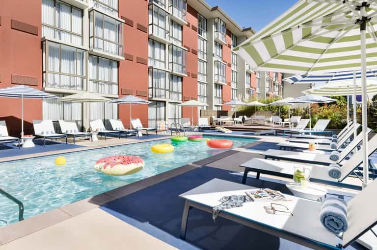 Modern resort pool with colorful floating pastry inflatables, striped umbrellas, and coral building