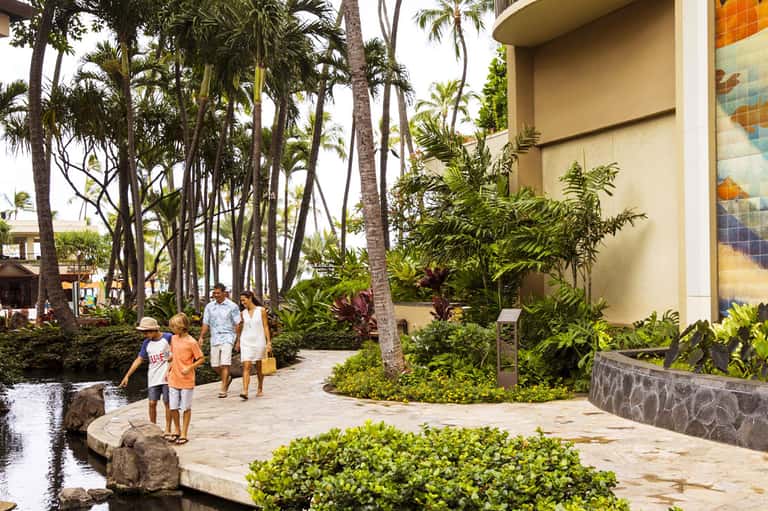 Family walking through tropical resort grounds with palm trees, water features, and lush landscaping