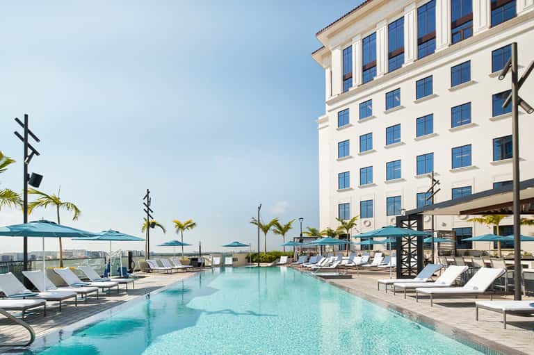 Resort infinity pool overlooking city skyline with white hotel building and palm trees