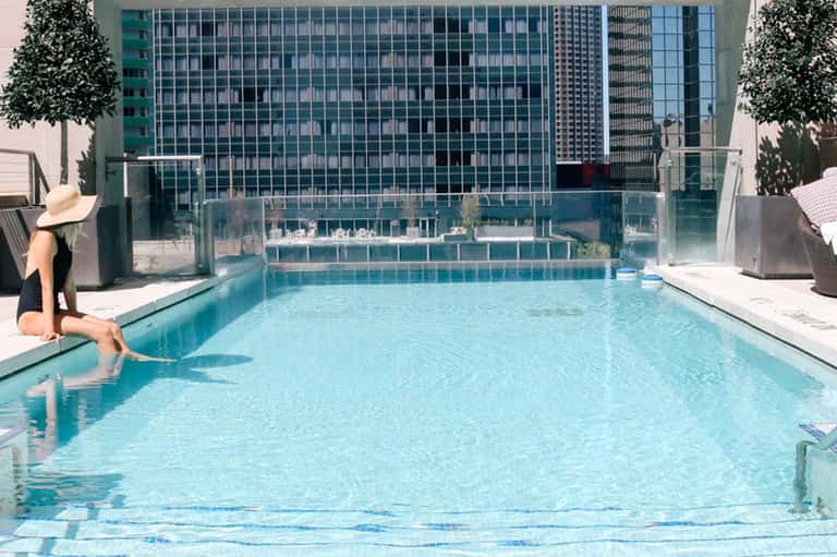 Urban rooftop pool with city skyline reflection, lounge chair, and surrounding high-rise buildings
