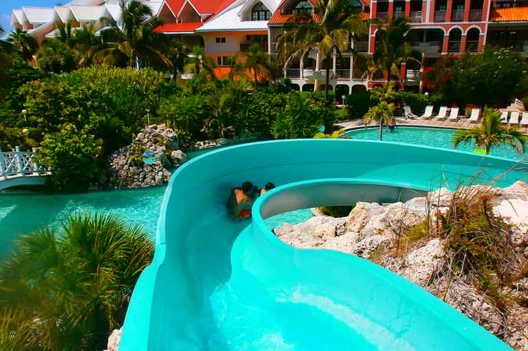 Blue water slide at resort with colorful buildings, palm trees, and lounge chairs
