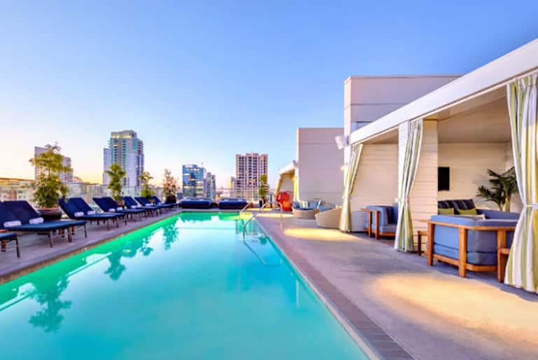 Rooftop pool with turquoise water overlooking city skyline at dusk, lounge chairs and cabanas