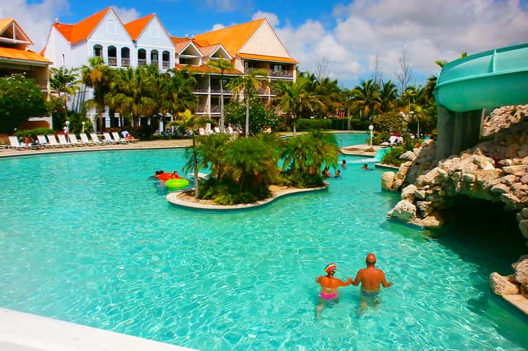 Resort pool with turquoise water, waterslide, and colonial-style orange-roofed buildings