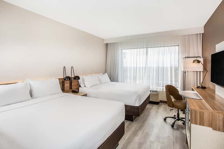 Contemporary hotel room with two queen beds, TV, and sheer curtains by window