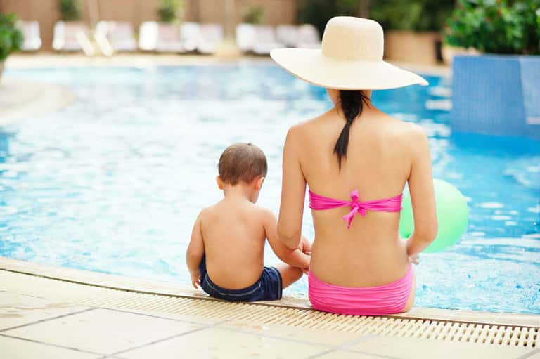 Woman and child sitting poolside at resort, child in water, woman wearing pink bikini and wide-brimmed hat