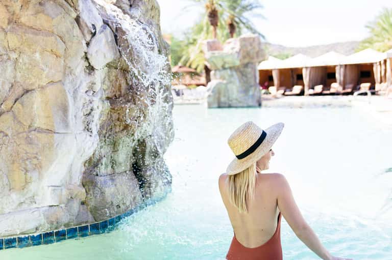 Woman in straw hat wading in turquoise thermal pool beside rocky cliff with resort loungers in background