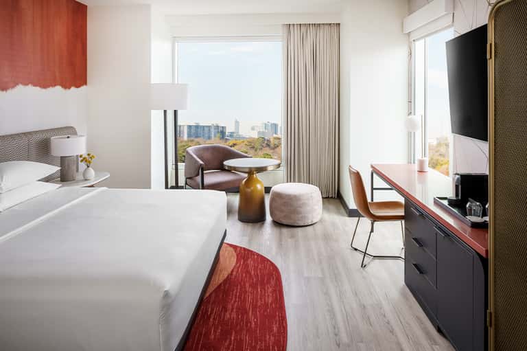 Modern hotel room with bed, seating area, cityscape view through floor-to-ceiling windows