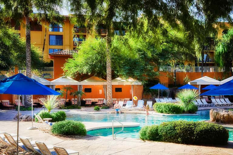 Resort pool area with blue umbrellas, lounge chairs, and orange building surrounded by palm trees and greenery
