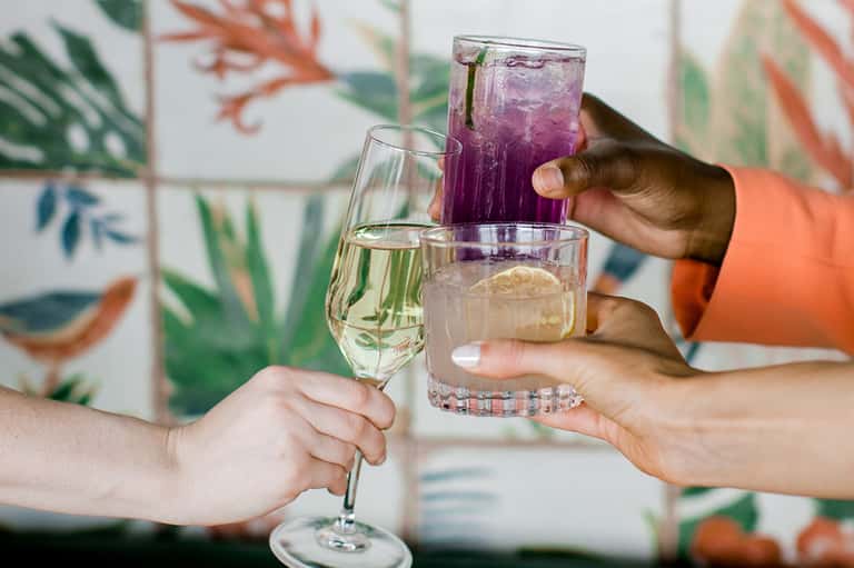 Three people clinking glasses with wine, water, and purple cocktail against a tropical botanical wallpaper backdrop