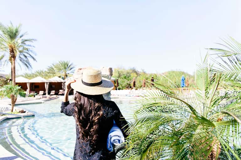 Person in straw hat viewing resort pool with palm trees and thatched-roof cabanas