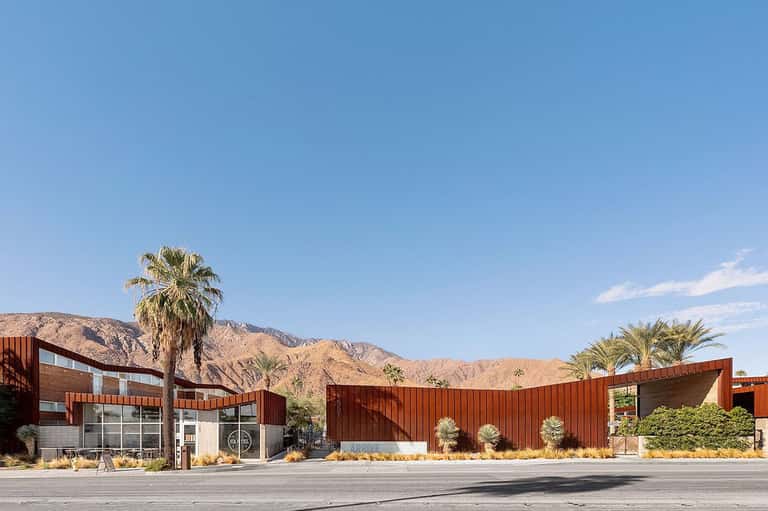 Modern resort building with corrugated metal facade and palm trees against desert mountains