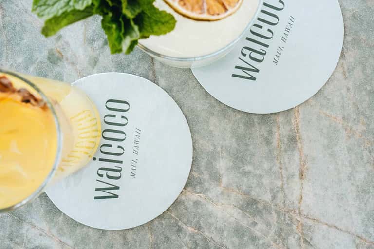 Two drinks on white coasters labeled 'wahcuu' with citrus garnishes on stone surface