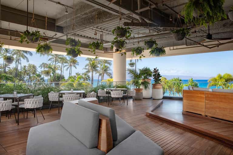 Waterfront lounge with wooden deck, hanging plants, modern furniture, and ocean view through glass railing