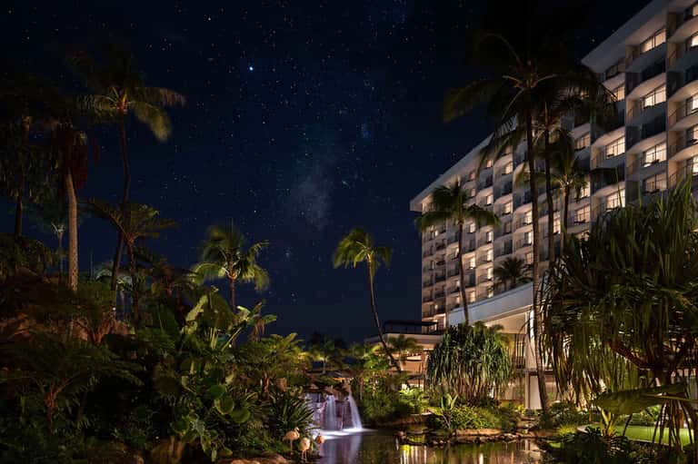 Nighttime resort view with illuminated hotel building, tropical plants, and waterfall