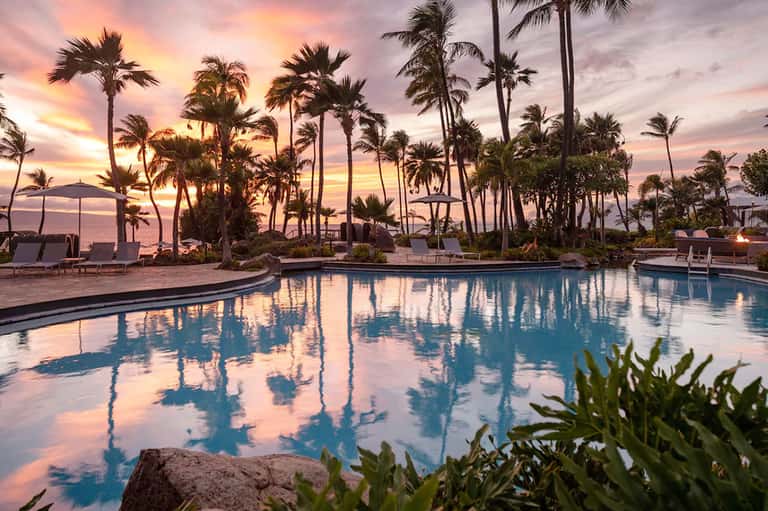 Resort pool at sunset with palm trees silhouetted against pink and purple sky, lounge chairs nearby
