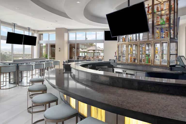 Modern hotel lobby bar with curved black counter, flat-screen TVs, and beverage display shelving