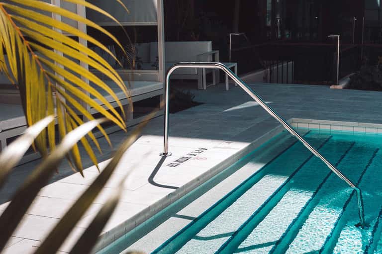Modern lap pool with stainless steel handrail and palm frond shadow, sunny day, hotel deck