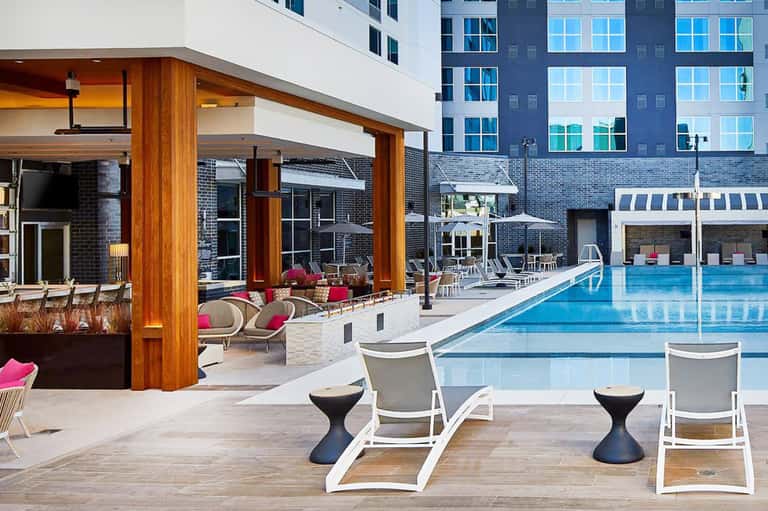 Resort pool area with lounge chairs, modern wooden pergola structure, pink accent furnishings, and multi-story hotel building backdrop