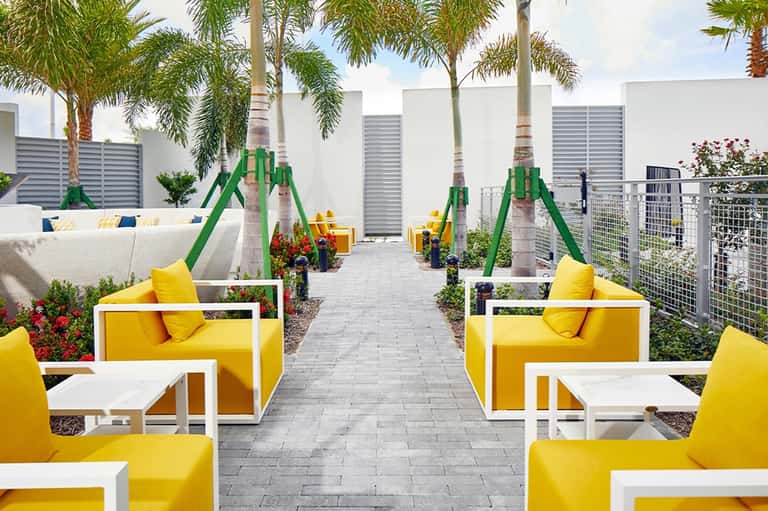 Modern outdoor lounge area with yellow chairs, white tables, palm trees, and green playground equipment