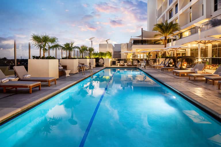 Illuminated resort rooftop pool at dusk with lounge chairs, fire pits, palm trees, and modern building architecture