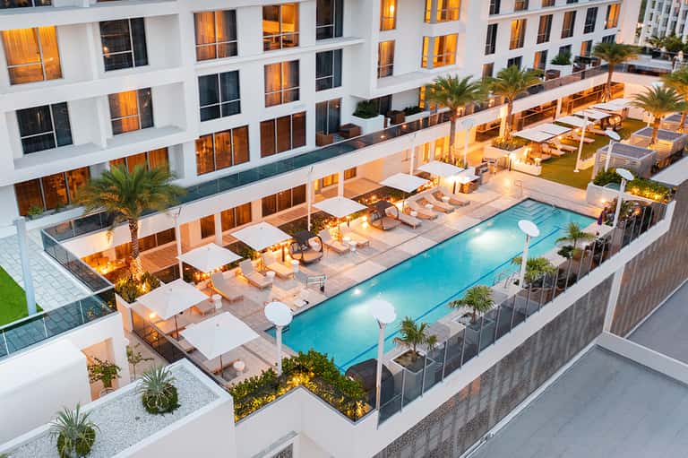 Aerial view of resort courtyard with illuminated pool, palm trees, lounge chairs, and white building facades