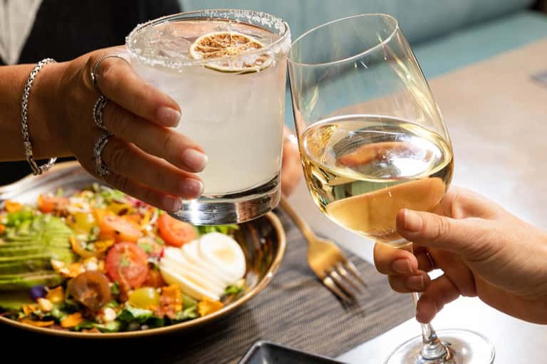 Two people toasting with cocktail and wine glass by pool with salad