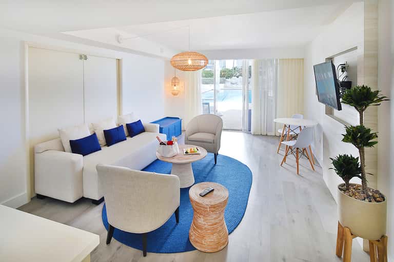 Modern hotel suite living area with white sofa, blue accents, and ocean view through sliding doors