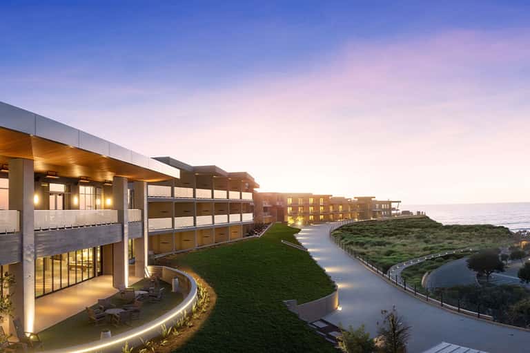 Modern curved resort buildings on grassy hillside at twilight with ocean view and lit pathways